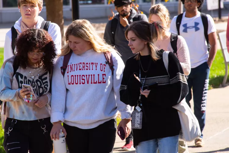 Group of female students walking on the sidewalk, female in the center wears a University of Louisville pullover.