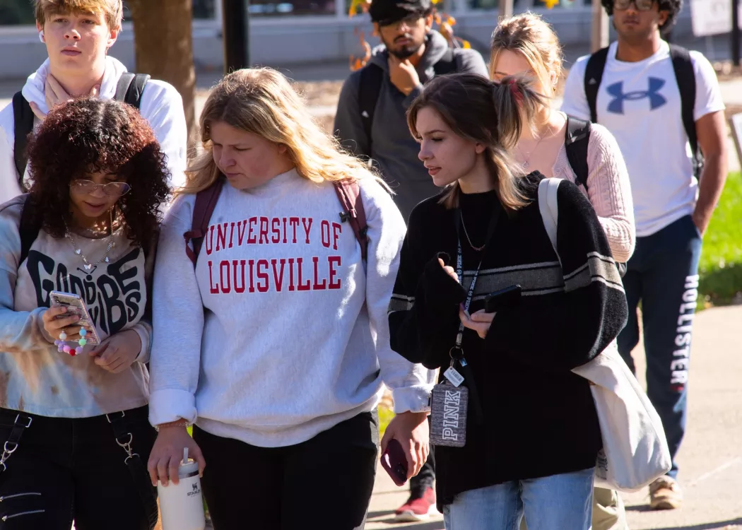 Group of female students walking on the sidewalk, female in the center wears a University of Louisville pullover.