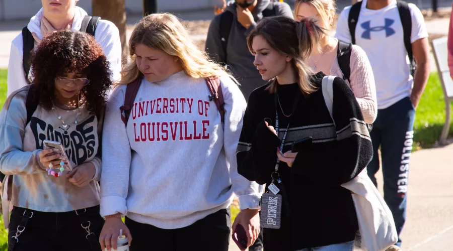 Group of female students walking on the sidewalk, female in the center wears a University of Louisville pullover.
