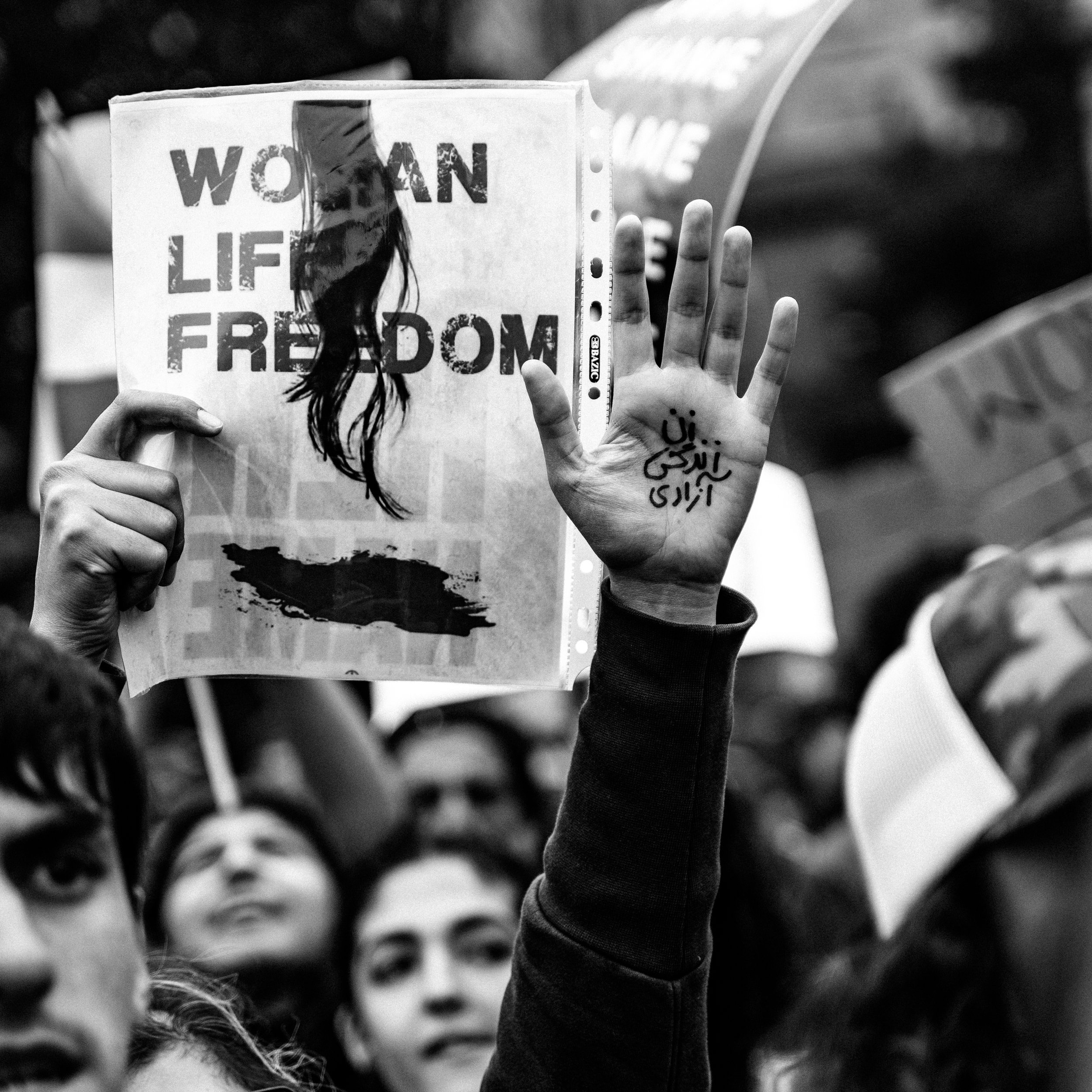 a black and white photograph by Chidi Nobi depicting a protest, one protestor has their hand raised with Sanskrit on their palm, another holds up a sign that reads women, life, freedom