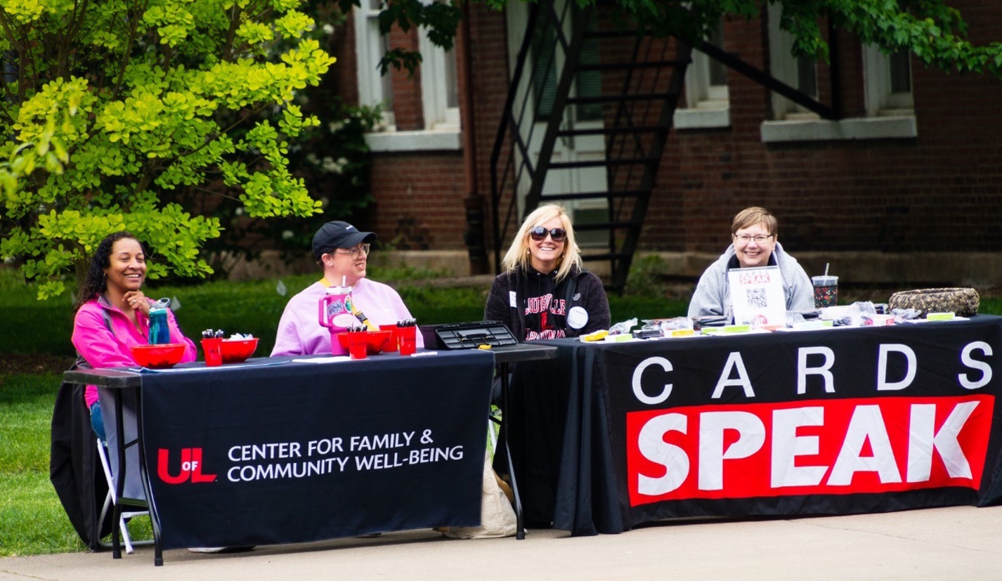 Photo of 4 people sitting and smiling at 2 covered tables. Banner on first table reads 'UofL Center for Family & Community Well-Being' and second table reads 'CARDS SPEAK'.