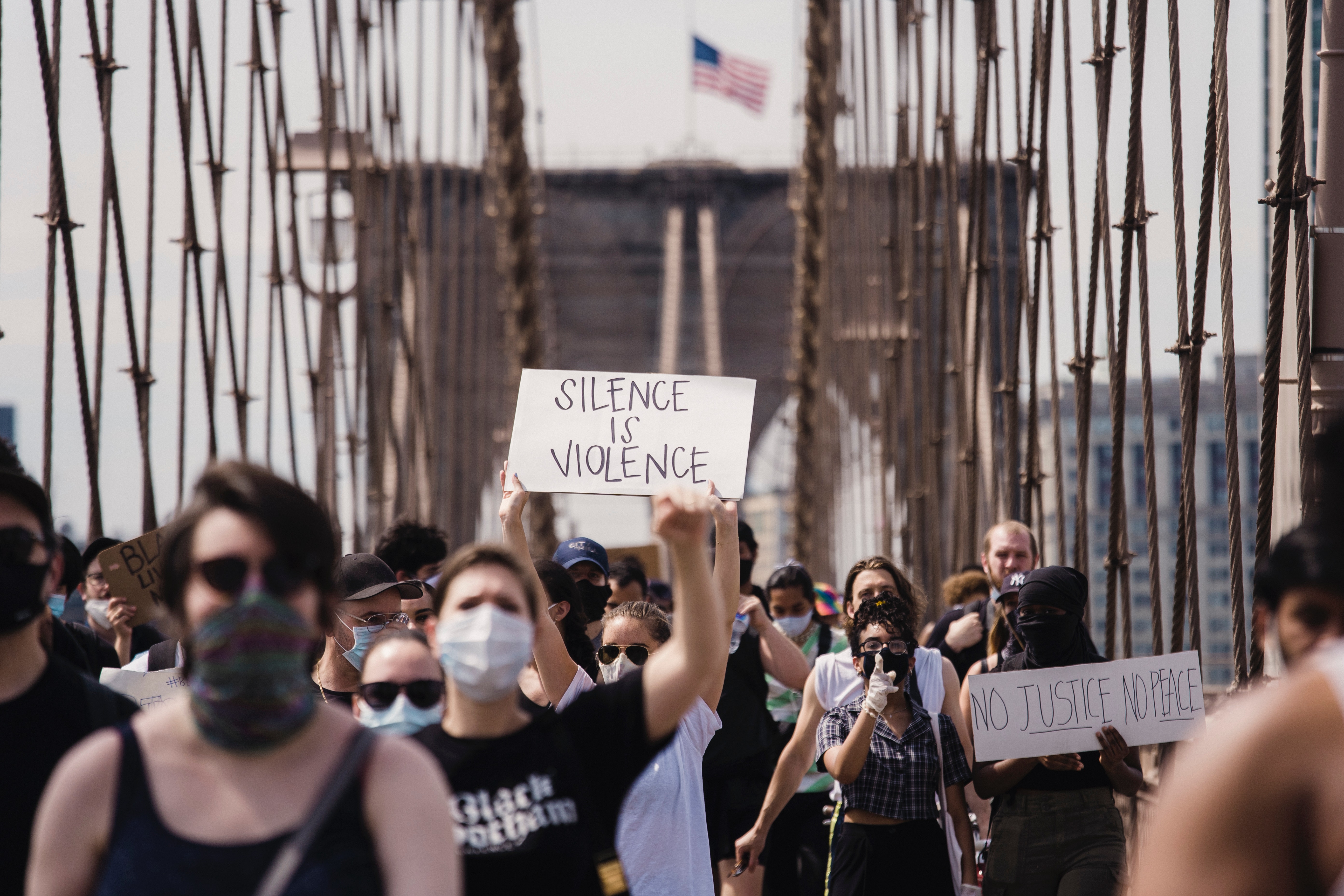 photo of peaceful protest on bridge with american flag in background and person in foreground holding sign that says 'Silence is Violence'