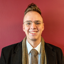 Gabriel Fiandeiro, a smiling white person with a brown mohawk and metal glasses, wears a brown pinstripe jacket, brown houndstooth scarf, and brown tie on a white button up shirt in front of a maroon background.