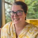Megan Heise, a white woman with brownish-red hair pulled back, glasses, and a yellow shirt, framed by the inside of an inclined plane car and beyond that, green trees.