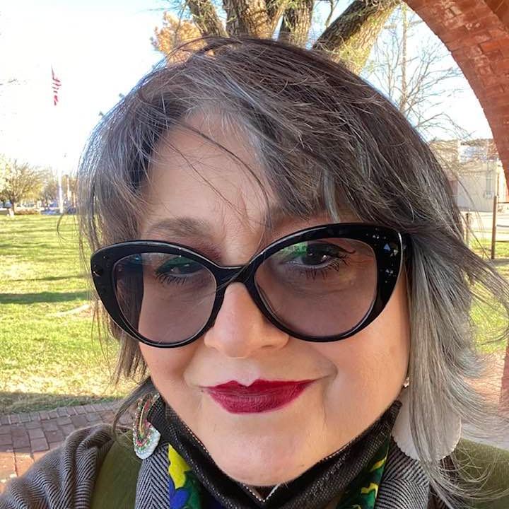 Kimberly Wieser-Weryackwe, a woman of medium complexion, stands outside on a sunny day in front of trees and brick arch, with her short gray hair blowing in the breeze, wearing large, black, cat eye glasses and white, pink, and green beaded oval earrings with mirrors in the center. 