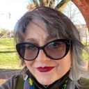 Kimberly Wieser-Weryackwe, a woman of medium complexion, stands outside on a sunny day in front of trees and brick arch, with her short gray hair blowing in the breeze, wearing large, black, cat eye glasses and white, pink, and green beaded oval earrings with mirrors in the center. 