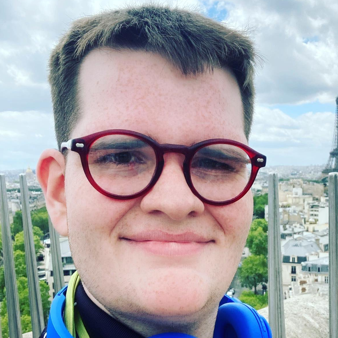 Headshot of young man with pale skin, brown hair, and wearing red glasses and blue headsets around his neck. He is standing in front of the Eiffel Tower. 