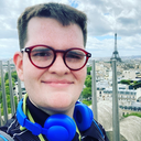 Headshot of young man with pale skin, brown hair, and wearing red glasses and blue headsets around his neck. He is standing in front of the Eiffel Tower.   