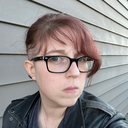 Aubrie Cox Warner, a white woman with red hair with shaved sides and black glasses. She is wearing a black leather jacket, blue shirt, and a bronze, pyramid-shaped pendant. Behind her is blue-gray siding.