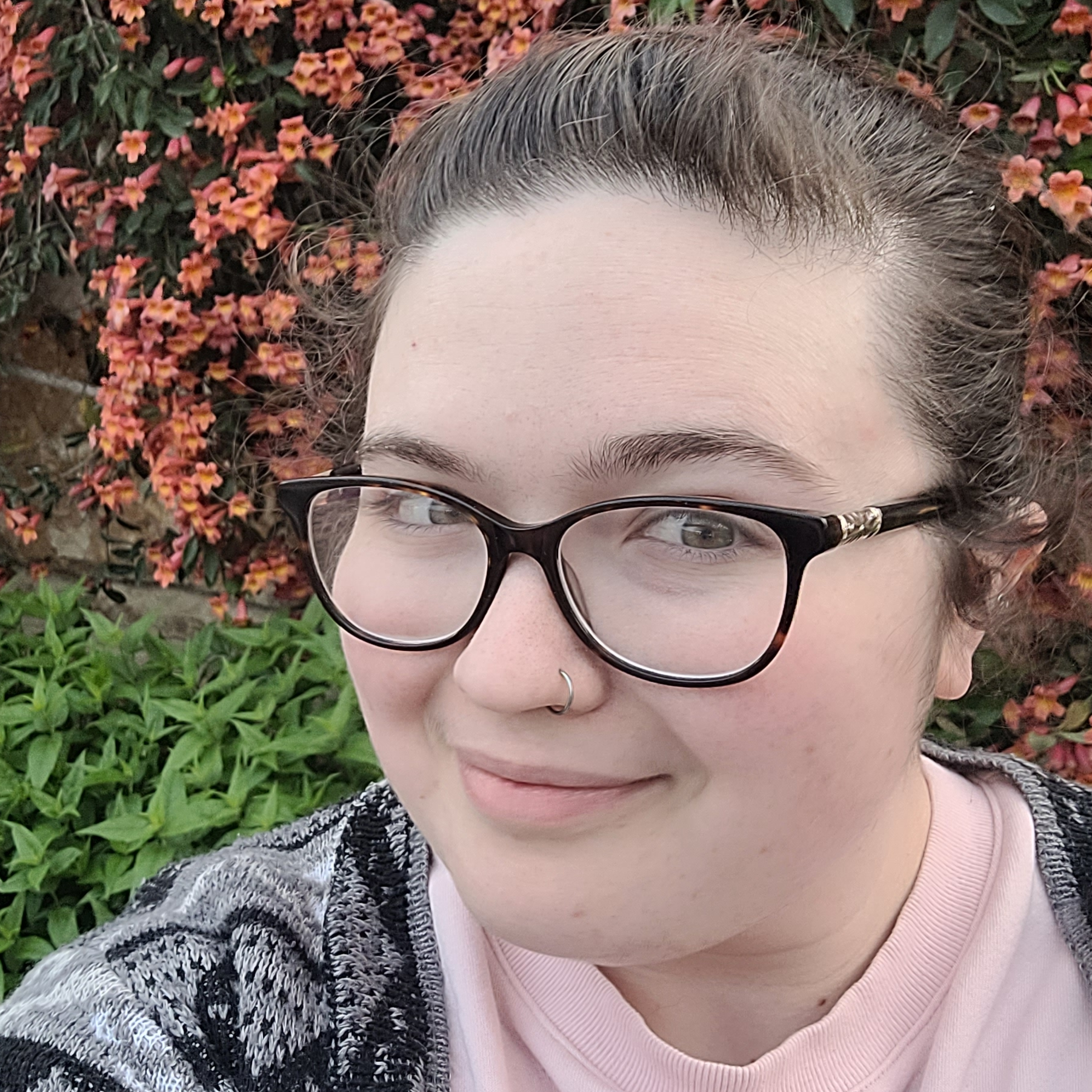 Kristen Wheaton, a smiling white woman with pulled back brown hair, a silver nose-ring and dark glasses. She is wearing a pink sweater and black and grey patterned cardigan while standing outside in front of a stone wall covered in red flowers above and green ivy below.