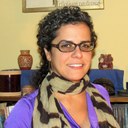 Anna Browne Ribeiro, a light-skinned Latinx woman with shoulder-length curly brown hair and glasses, is framed in front of red and white ceramic objects Anna T. Browne Ribeiro