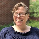 Photo of a woman in glasses smiling, a brick wall and plants in the background.  Carrie Mott