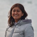 Hilaria Cruz, a brownskin woman with brown hair who is smiling. She is standing against a white background. Hilaria Cruz
