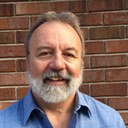 Michael Pemberton, a white male with a trimmed grey beard, in front of a brick wall. Michael Pemberton