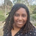 Michelle Grue, a Black woman with medium length locs, standing in front of trees. Michelle Grue