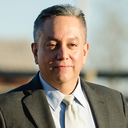 Dr. Ron Sheffield, a Native American man with short salt-and-pepper hair color, is framed wearing a charcoal-colored suit with a light yellow-shaded tie. Ron Sheffield