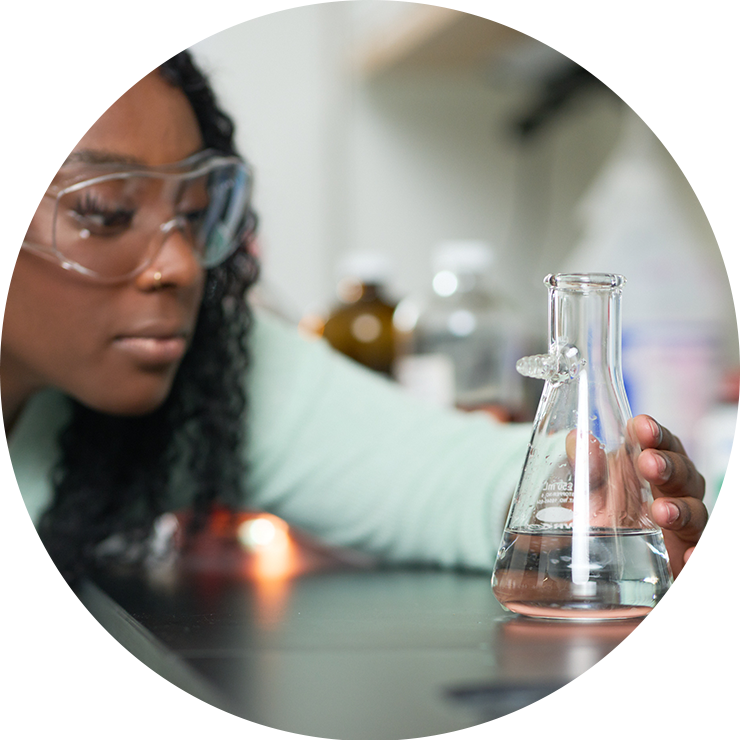 Close-up photo of a female undergraduate student wearing safety glasses investigating a chemical reaction in a graduated flask in a chemistry lab.
