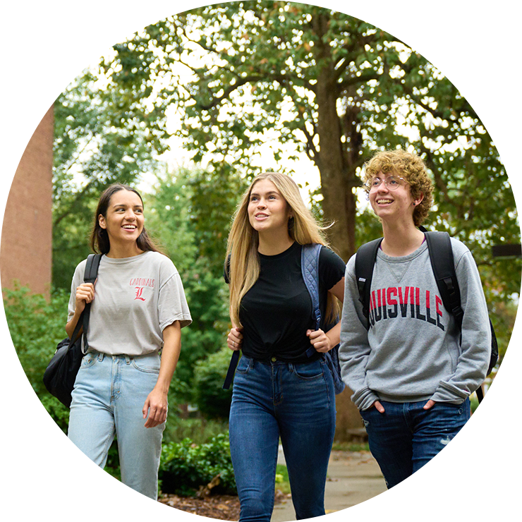 Three students walking across campus, talking and smiling on an overcast day, with an academic building and trees in the background.