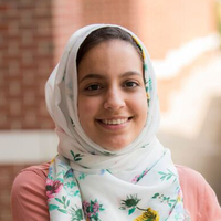 Rianne Kablan; a scholar wearing a pink shirt and white floral headscarf standing outside in front of a brick wall.