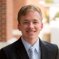 Ryan Crum; a scholar wearing a black suit and blue tie standing outside in front of a brick wall.