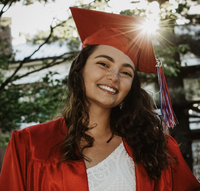 An image of a young woman of color wearing a red graduation cap and gown. She has long, wavy hair. She is standing outside in a wooded area with the setting sun behind her.
