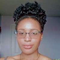 A headshot of a young woman of color wearing glasses. She is indoors. Her hair is curly and gathered into a updo. 