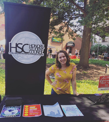 An image of Allison LaRoy, a student in a yellow floral shirt, standing outside at a table. Behind the table is a black banner with an owl logo that reads 
