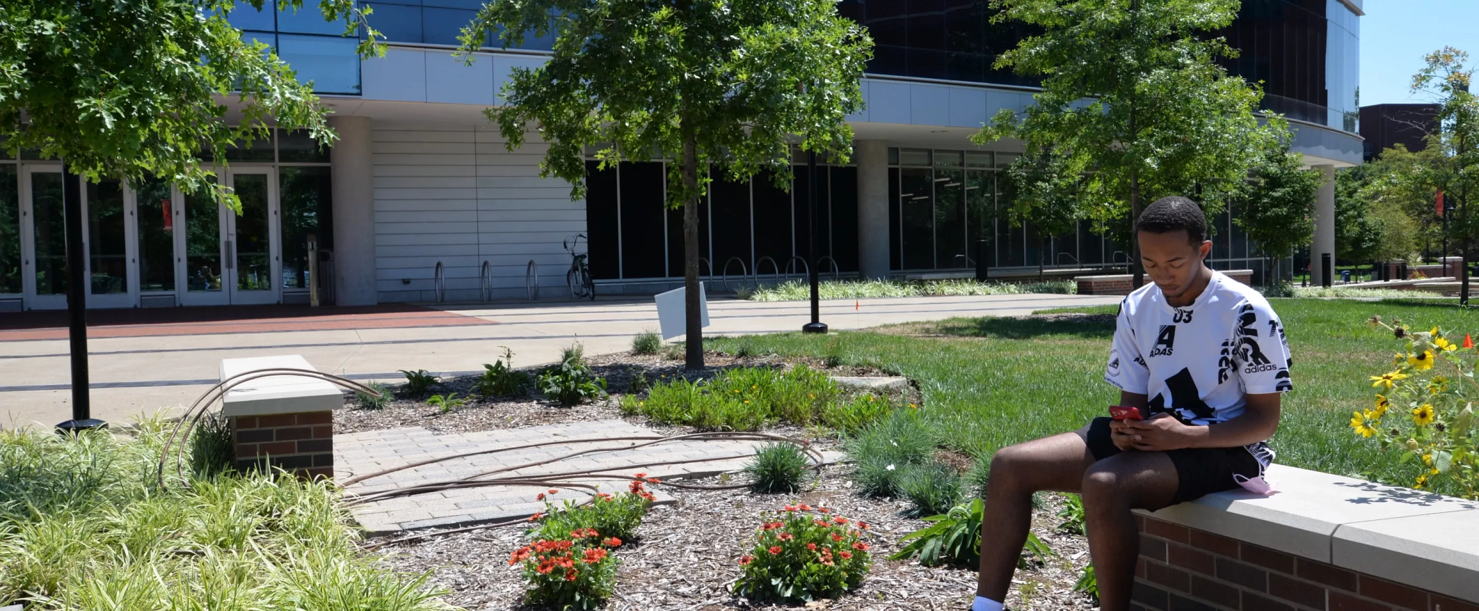 Male student sitting on bench outside of the Belknap Academic building. He's looking at his phone.