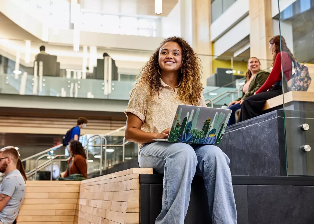 Student recognizes another student walk into the Belknap Academic Building as she is typing on her laptop and sitting on the wooden platform benches at the entrance of the BAB.