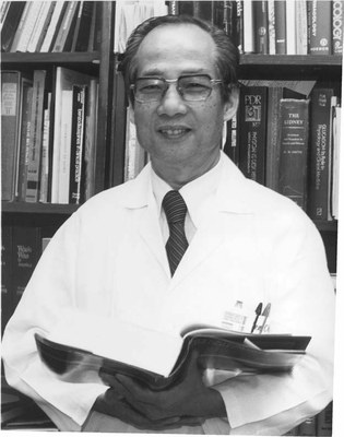 KC Huang, dressed in labcoat, stands in front of book case and holds a book open.