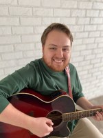 Andrew Chapman, MT-BC, is the coordinator of the UofL MT Clinic. He is pictured smiling with a green shirt and burgundy burst acoustic guitar in front of a white brick wall.
