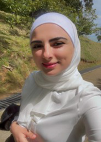 A smiling woman in a white hijab and white dress stands in front of a grassy hill