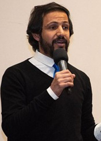 A man with dark hair and a beard wearing a shirt and dark sweater speaks into a microphone