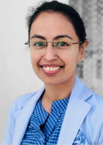 A smiling woman with dark hair pulled back and glasses wearing a patterned blue blouse and a light blue blazer stands before a grey background