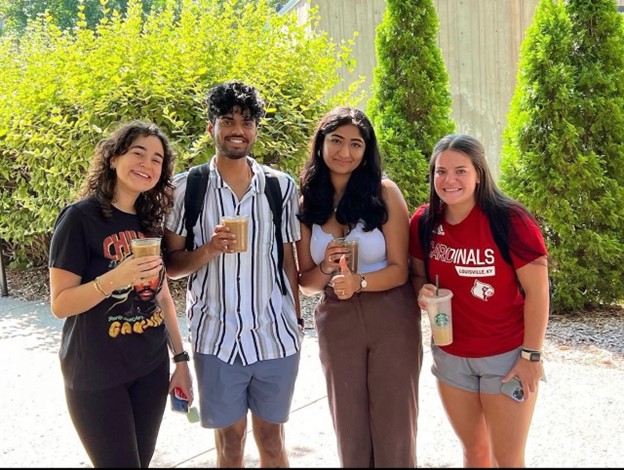 Four students posing with coffee outside in front of bushes.