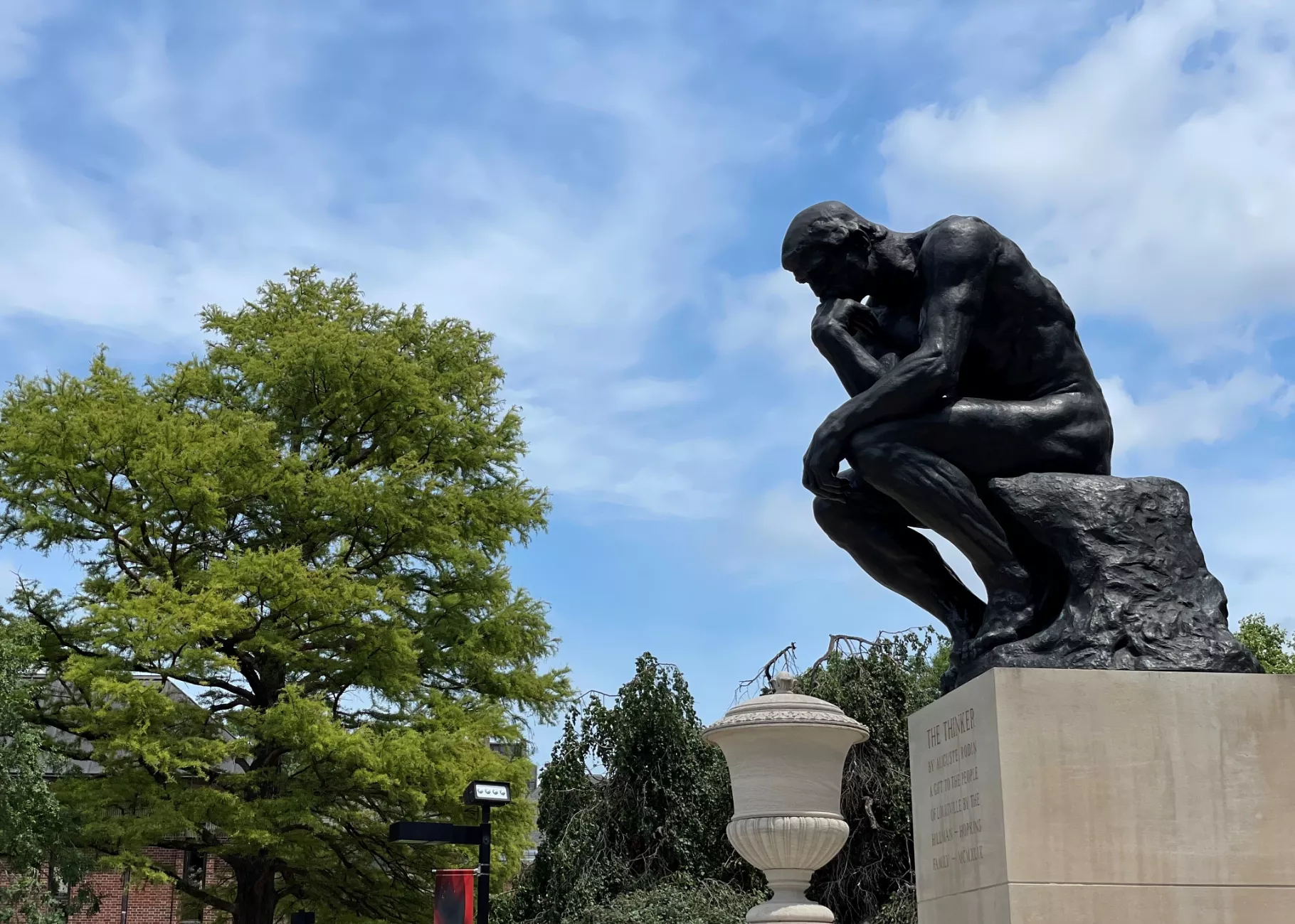 UofL's thinker statue from the side with the sky in the background. Trees on campus can be seen in the background as well as…