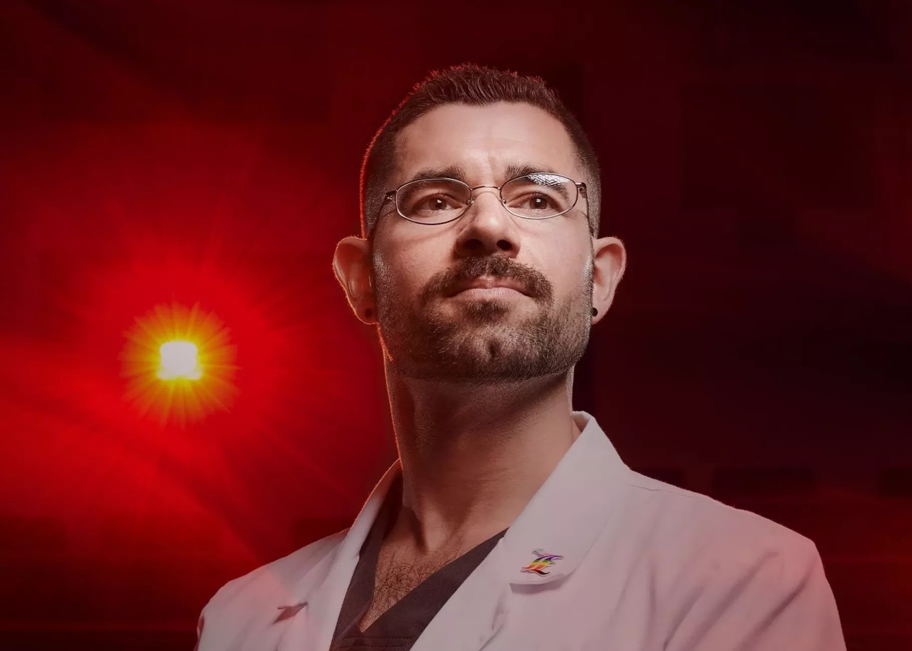 A man with glasses in a white lab coat stands in front of a red spotlight.