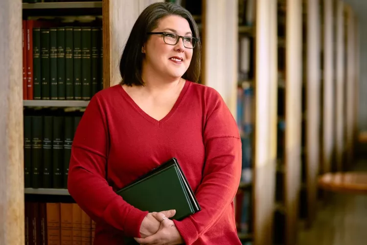 A smiling woman with glasses and holding books stands among shelves of library books.