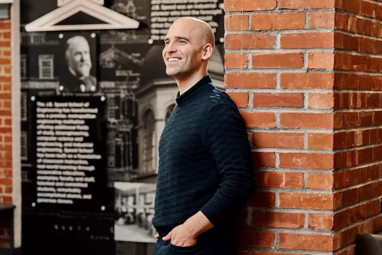 A smiling man in a black sweater poses against an industrial brick wall.