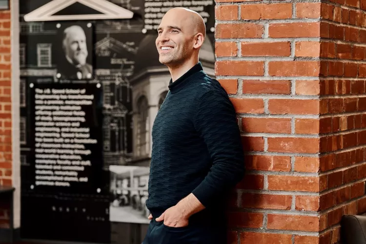 A smiling man in a black sweater poses against an industrial brick wall.