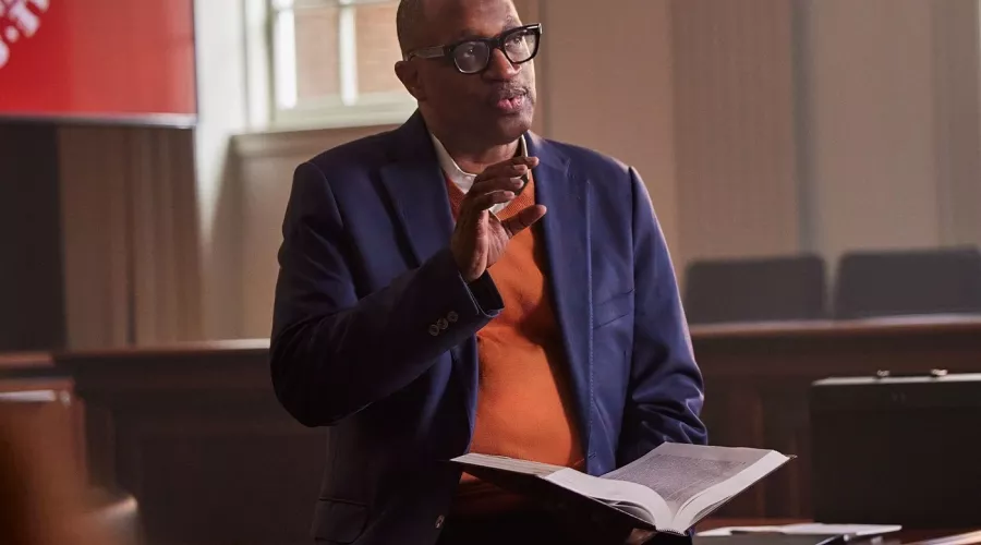 A professor with glasses and holding a book lectures in a school courtroom.