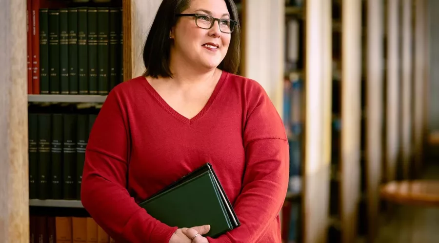 A smiling woman with glasses and holding books stands among shelves of library books.