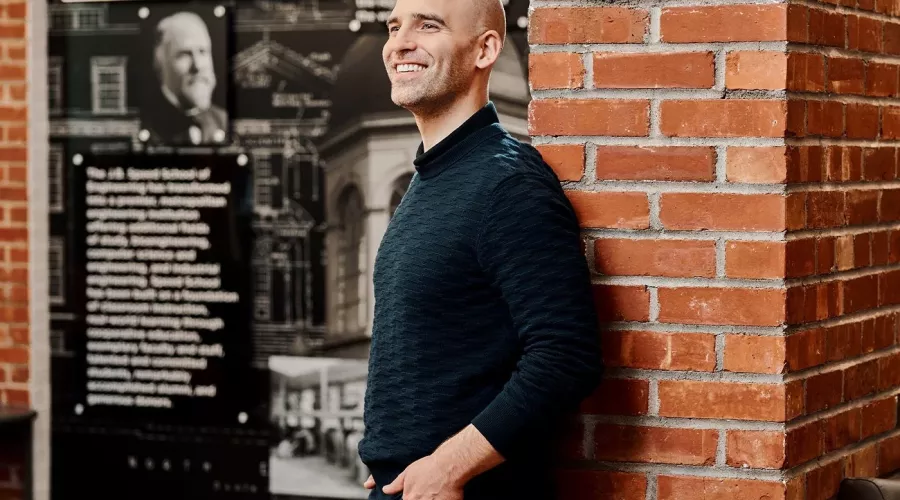 A smiling man in a black sweater poses against an industrial brick wall.