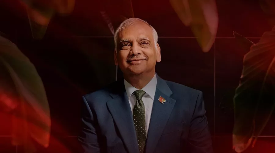 A smiling man in a dark suit sits on a wooden bench surrounded by palm leaves.