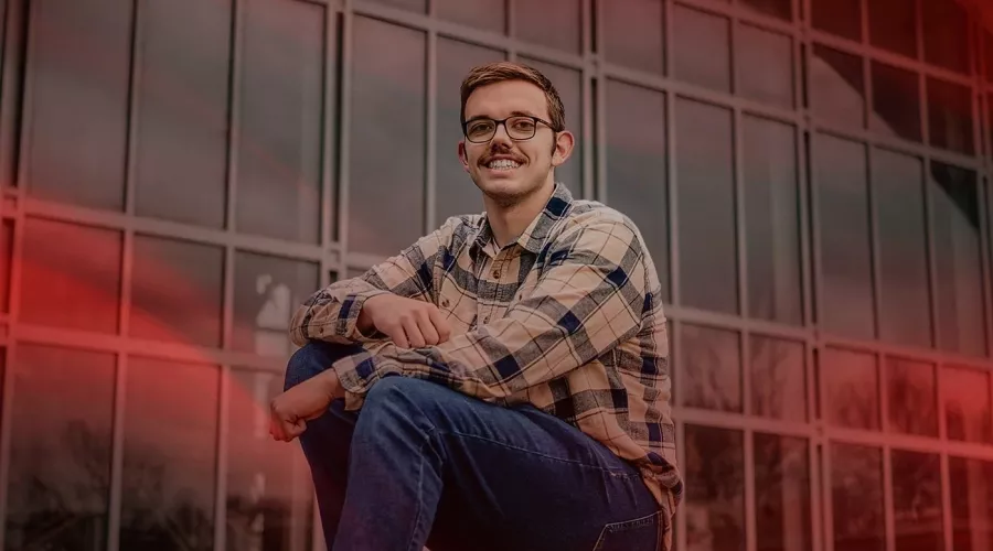 A smiling man with glasses sits on outdoor steps in front of a glass-front building.