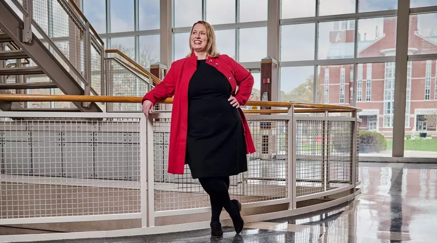 A smiling woman in a red jacket and black dress poses against a stairway railing.