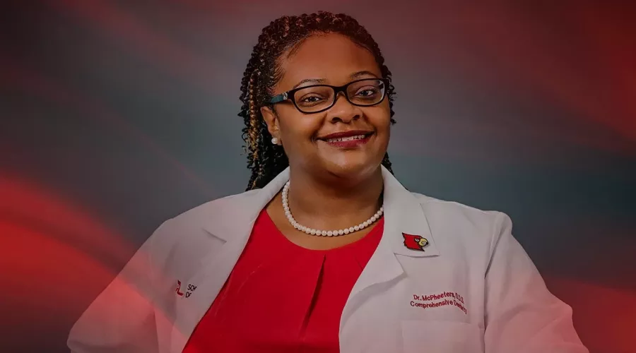 A smiling woman with glasses in a white lab coat leans on a dentist's chair.