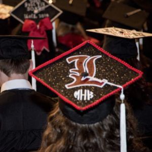 Graduates in caps facing away from the viewer, closest has a large L on the top.