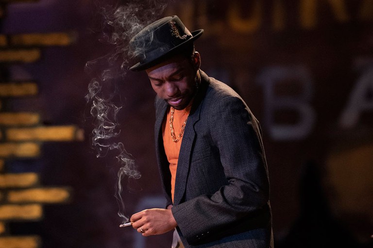 A man in a fedora and suit jacket smokes a cigarette 