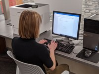 the back of a woman with blonde hair sitting at a chair facing a computer and typing